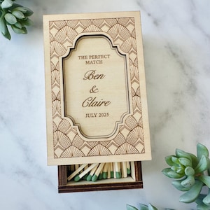 Può includere: Un portasigarette in legno con un cassetto che scorre per rivelare fiammiferi. La scatola è decorata con un design art déco e il testo "THE PERFECT MATCH Ben & Claire JULY 2025".