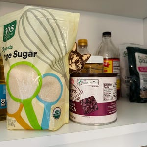 May include: A white bag of 365 Everyday Value Organic Cane Sugar, a can of Ghirardelli Unsweetened Cocoa, and other pantry items on a white shelf.