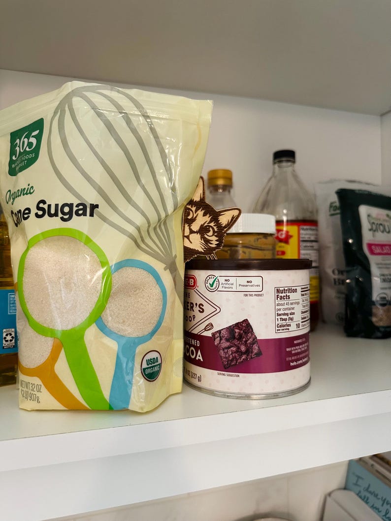 May include: A white bag of 365 Everyday Value Organic Cane Sugar, a can of Ghirardelli Unsweetened Cocoa, and other pantry items on a white shelf.