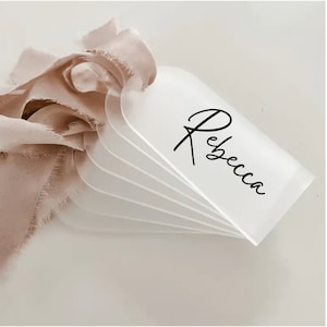 May include: A stack of frosted acrylic place cards with rounded tops, each bearing the name "Rebecca" in elegant black script. A blush-colored ribbon is attached to the cards, adding a soft, feminine touch.