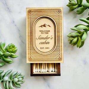Pu&ograve; includere: Scatola di fiammiferi in legno con la scritta "WELCOME TO THE Sander's cabin EST. 2001" e un grafico di montagna. La scatola &egrave; aperta, rivelando fiammiferi con punte bianche. Il design &egrave; inciso nel legno. Succulente verdi sullo sfondo.