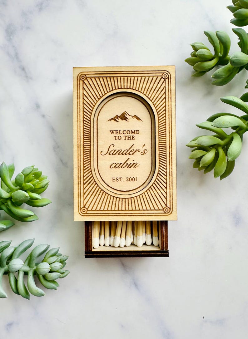 Pu&ograve; includere: Scatola di fiammiferi in legno con la scritta "WELCOME TO THE Sander's cabin EST. 2001" e un grafico di montagna. La scatola &egrave; aperta, rivelando fiammiferi con punte bianche. Il design &egrave; inciso nel legno. Succulente verdi sullo sfondo.