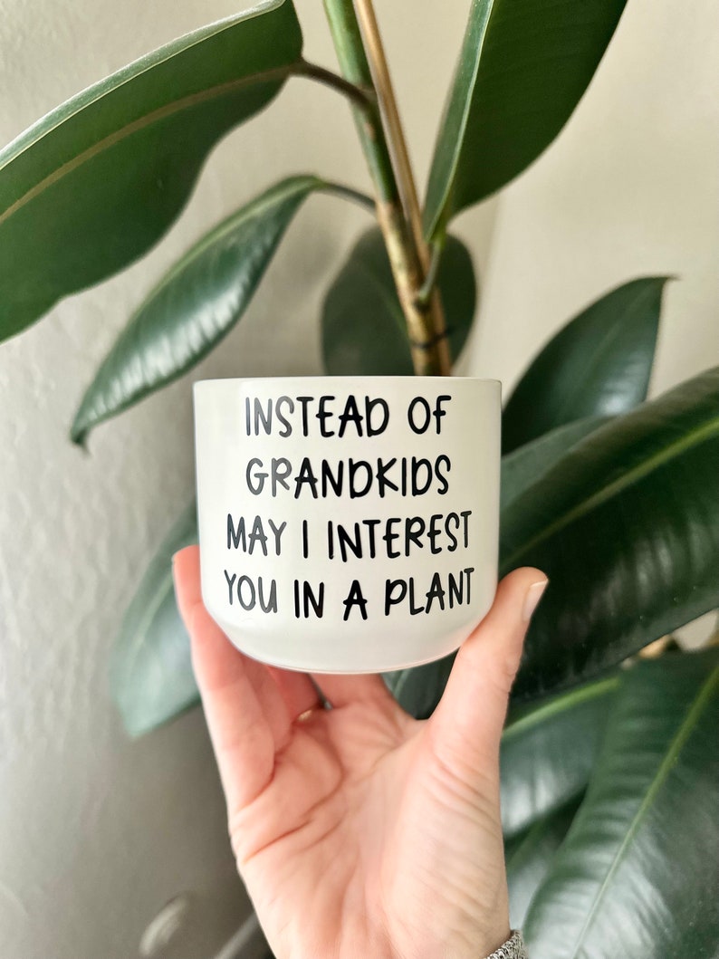 May include: White ceramic planter with black text that reads "Instead of Grandkids May I Interest You in a Plant."