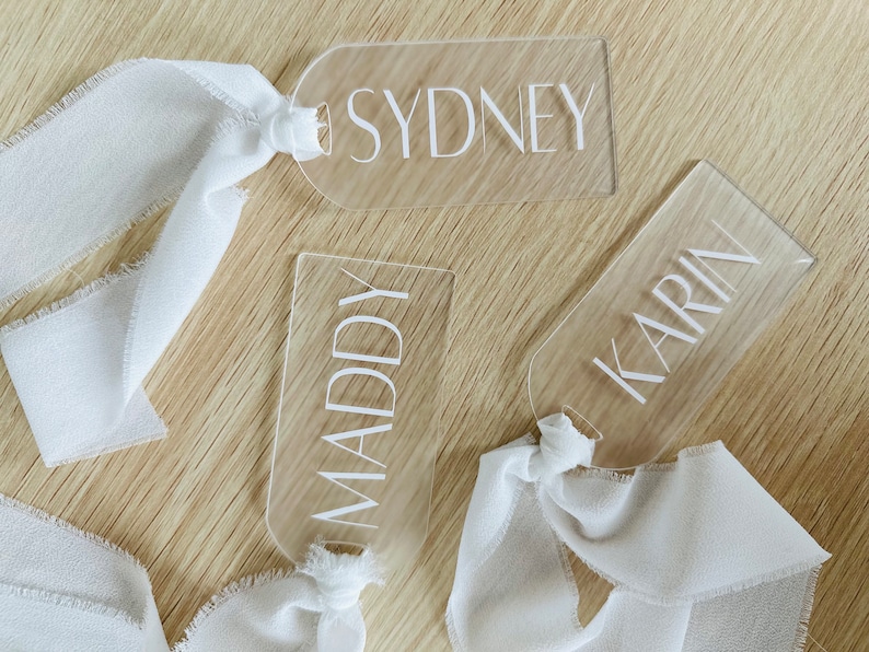 May include: Three clear acrylic luggage tags with white text. The tags are tied with white ribbon and the names "Sydney", "Maddy", and "Karin" are printed on them.