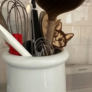 May include: A white ceramic utensil holder with a variety of kitchen tools inside, including a wooden spoon, a whisk, and a pair of tongs. A wooden cat head cutout is peeking out from behind the tools.