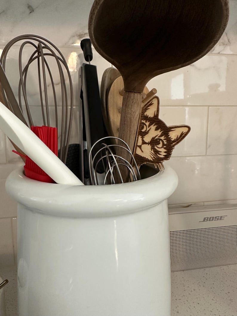 May include: A white ceramic utensil holder with a variety of kitchen tools inside, including a wooden spoon, a whisk, and a pair of tongs. A wooden cat head cutout is peeking out from behind the tools.
