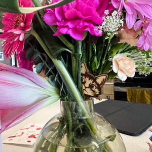May include: A bouquet of pink, white, and peach flowers in a clear glass vase. A wooden cat figurine is tucked behind the stems.