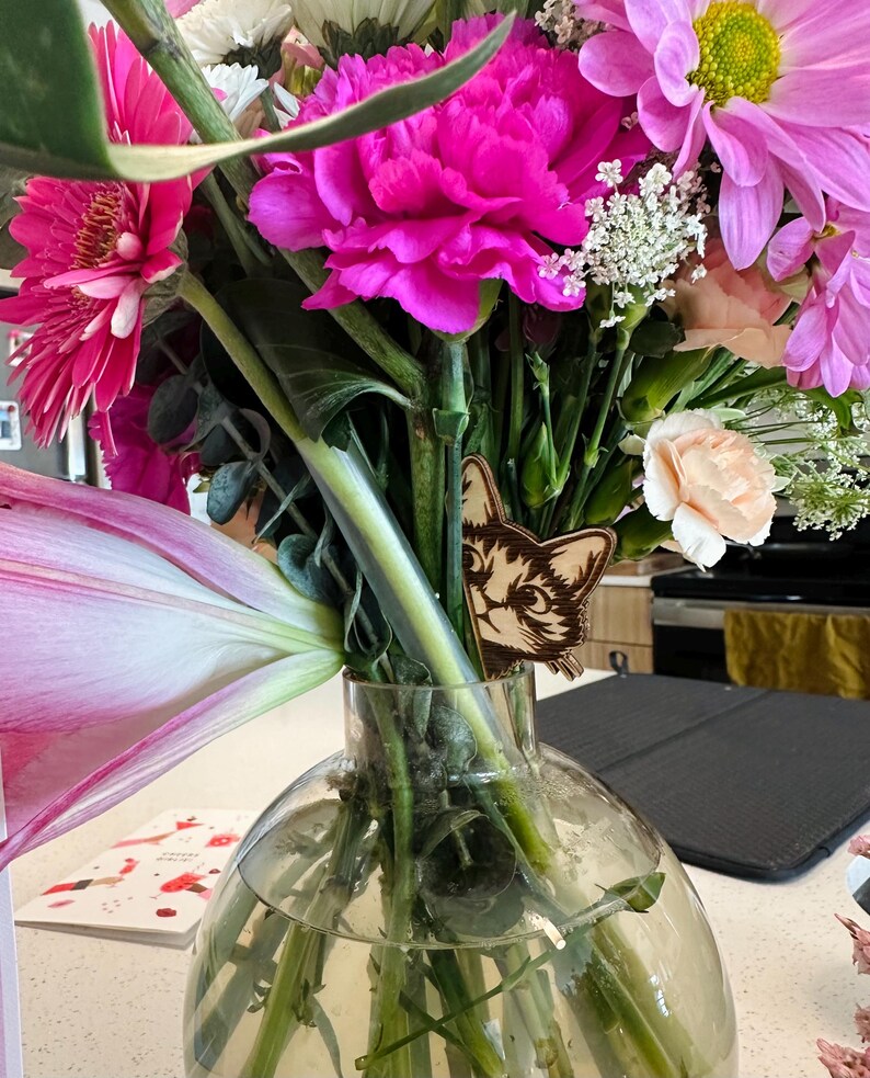 May include: A bouquet of pink, white, and peach flowers in a clear glass vase. A wooden cat figurine is tucked behind the stems.