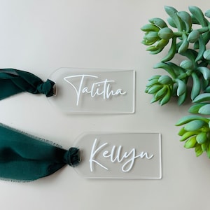 May include: Two clear acrylic place cards with the names "Talitha" and "Kellyn" written in white cursive script. Each card is tied with a green ribbon.
