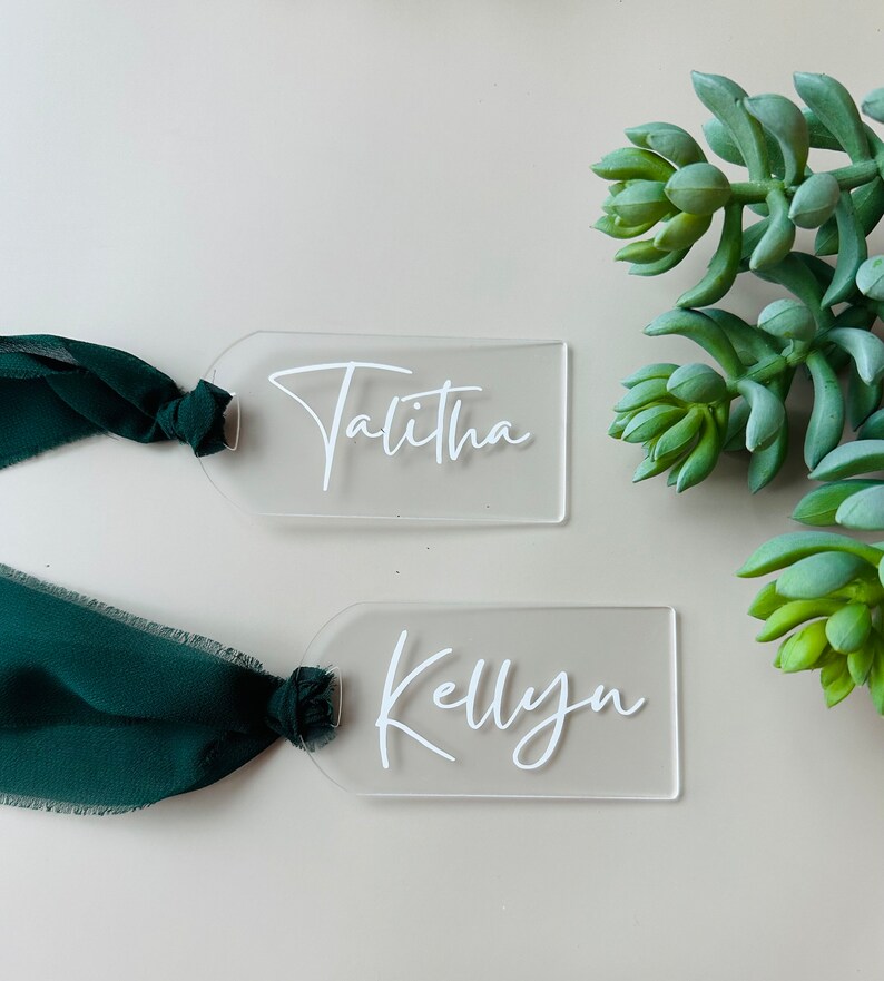 May include: Two clear acrylic place cards with the names "Talitha" and "Kellyn" written in white cursive script. Each card is tied with a green ribbon.
