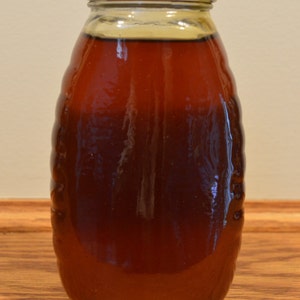 May include: A clear glass bottle with a gold lid filled with a dark brown liquid. The bottle has a textured surface and is sitting on a wooden surface.