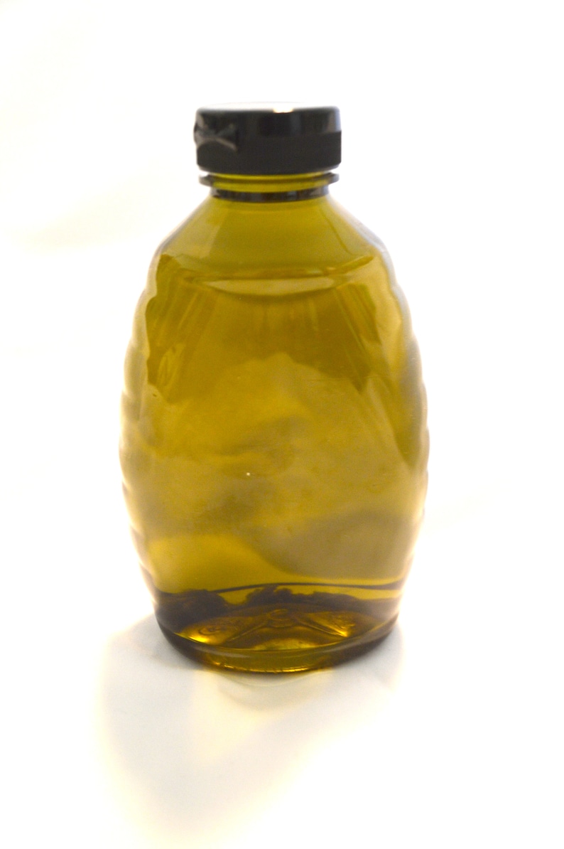 May include: A clear glass bottle with a dark brown, textured surface. The bottle has a black lid and is filled with a light brown liquid.