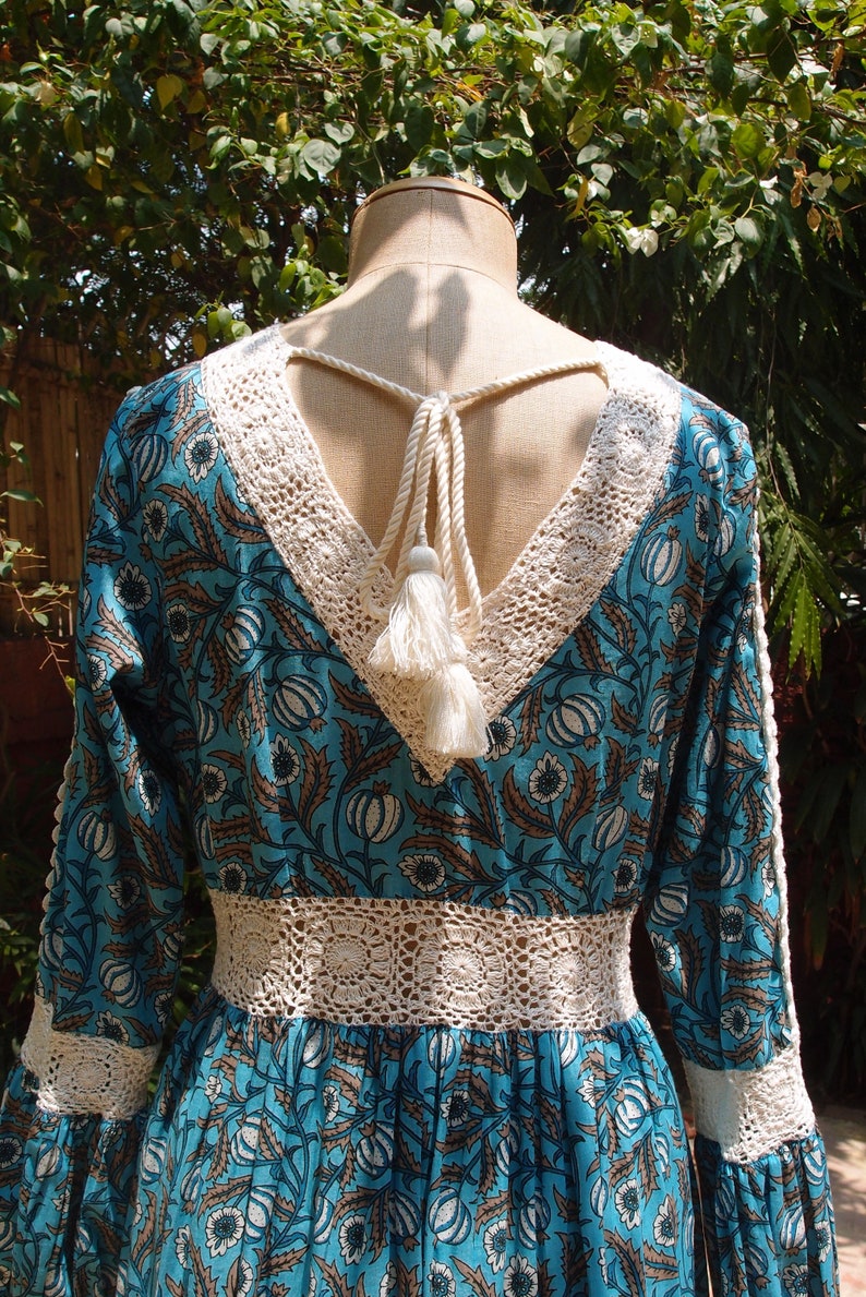Pode incluir: Um vestido maxi de estampa floral azul e branco com acabamento em renda de croch&ecirc; e um la&ccedil;o de franja branco nas costas.