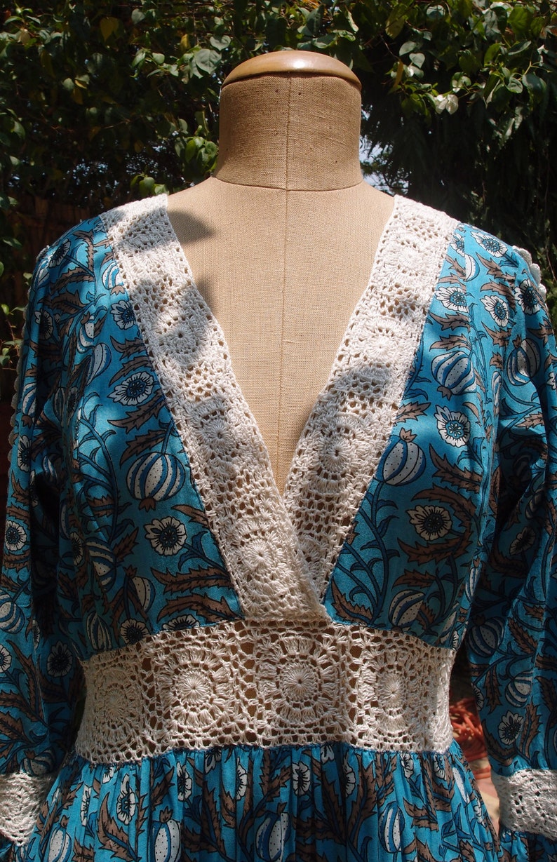 Pode incluir: Um vestido floral azul e branco com uma borda de croch&ecirc; em torno do decote e da cintura. O vestido tem um decote em V e mangas compridas.