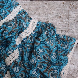 Pode incluir: Tecido azul com um padr&atilde;o floral branco e uma borda de croch&ecirc; branca. O tecido est&aacute; dobrado e drapeado sobre uma superf&iacute;cie de madeira clara.