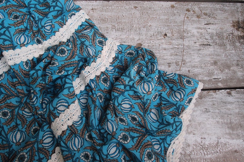 Pode incluir: Tecido azul com um padr&atilde;o floral branco e uma borda de croch&ecirc; branca. O tecido est&aacute; dobrado e drapeado sobre uma superf&iacute;cie de madeira clara.