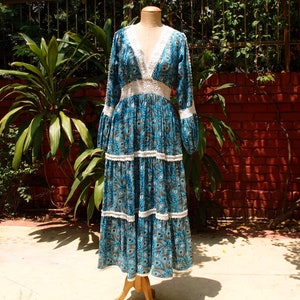 Pode incluir: Um vestido maxi longo floral azul e branco com decote e guarni&ccedil;&atilde;o de renda branca. O vestido tem uma saia em camadas e mangas compridas.