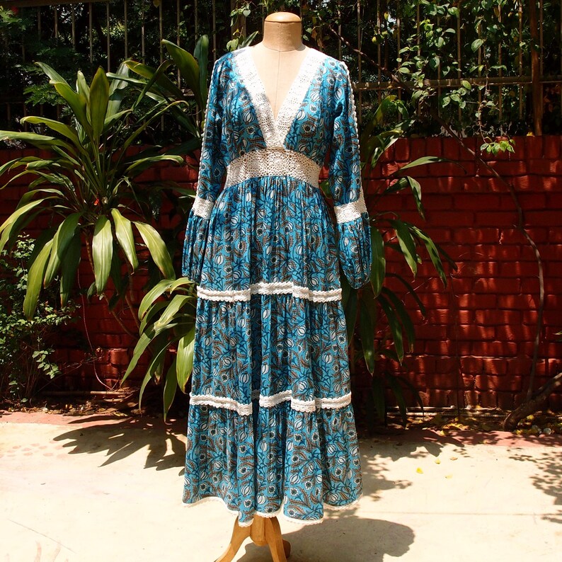 Pode incluir: Um vestido maxi longo floral azul e branco com decote e guarni&ccedil;&atilde;o de renda branca. O vestido tem uma saia em camadas e mangas compridas.