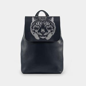 Könnte beinhalten: Ein marineblauer Rucksack mit Klappenverschluss und Tragegriff. Die Klappe zeigt eine detaillierte silberfarbene Illustration eines Luchskopfes. Der Rucksack hat ein schlichtes, stromlinienförmiges Design.
