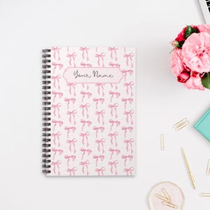 Puede incluir: Una libreta de espiral blanca con un patrón de lazos rosas y el texto "Your Name" en la portada.