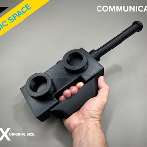 Op de afbeelding: Een zwarte plastic replica van een Star Trek-communicator, met een handvat en een lange antenne. De tekst "COMMUNICATOR" is gedrukt op de bovenkant van het apparaat. Het apparaat wordt in de hand van een persoon gehouden. De tekst "13X ORIGINAL SIZE" is gedrukt onder het apparaat.