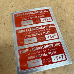May include: Three silver and red labels with the text "TORR LABORATORIES, INC. LOS ANGELES, CALIFORNIA HIGH VOLTAGE RELAY" and the type and serial number of the relay. The serial numbers are 4911, 4885, and 2967.