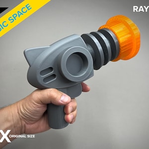 May include: A gray plastic toy ray gun with an orange plastic barrel. The gun is 13 times the original size. The text "RAYGUN" is visible in the upper right corner of the image.