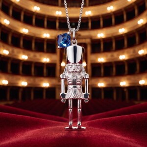 May include: A silver-toned nutcracker pendant hanging from a delicate box chain necklace. The pendant features a detailed soldier figure with a tall hat and a small, faceted deep blue crystal bead attached to the top ring near the chain bail.