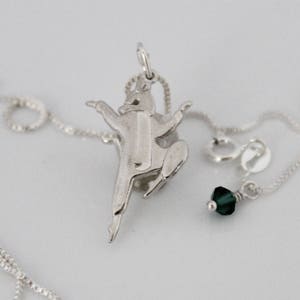 May include: A silver necklace with a pendant of a stylized figure in a martial arts pose. The necklace also has a small, dark green crystal charm.