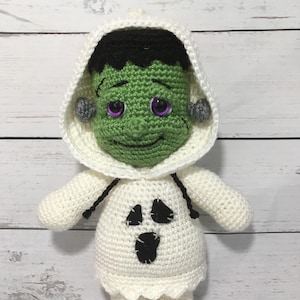 May include: A crocheted plush toy dressed as a ghost with a Frankenstein face. The ghost is white with black eyes and a black mouth. The Frankenstein face is green with black hair and purple eyes.