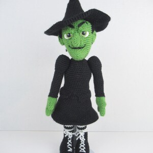 Puede incluir: Una mu&ntilde;eca de bruja de crochet verde con cabello negro, un sombrero negro y un vestido negro. La mu&ntilde;eca lleva guantes verdes y botas rayadas en blanco y negro. La mu&ntilde;eca est&aacute; de pie sobre un soporte blanco.