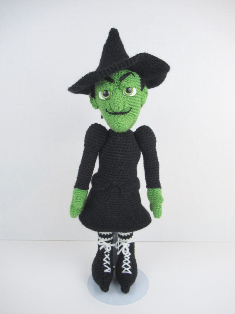 Puede incluir: Una mu&ntilde;eca de bruja de crochet verde con cabello negro, un sombrero negro y un vestido negro. La mu&ntilde;eca lleva guantes verdes y botas rayadas en blanco y negro. La mu&ntilde;eca est&aacute; de pie sobre un soporte blanco.