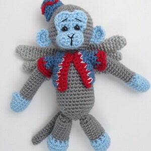 Puede incluir: Un mono de crochet gris que lleva un chaleco azul y rojo y un sombrero azul. El mono tiene ojos azules y una cara azul. El mono tiene alas y est&aacute; sentado sobre un fondo blanco.