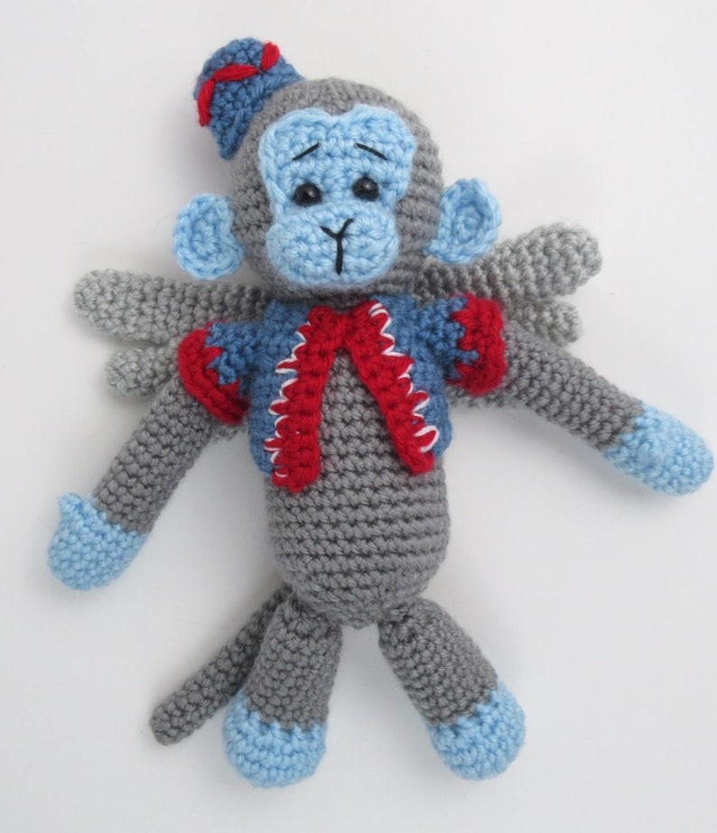 Puede incluir: Un mono de crochet gris que lleva un chaleco azul y rojo y un sombrero azul. El mono tiene ojos azules y una cara azul. El mono tiene alas y est&aacute; sentado sobre un fondo blanco.