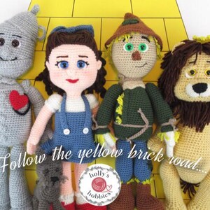 Op de afbeelding: Gehaakte pluche speelgoed van Dorothy, de Vogelsverschrikker, de Tin Man en de Laffe Leeuw uit The Wizard of Oz. De speelgoedfiguren staan op een gele bakstenen weg met de tekst "Follow the yellow brick road..." eronder.