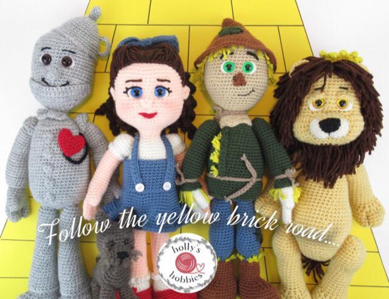 Op de afbeelding: Gehaakte pluche speelgoed van Dorothy, de Vogelsverschrikker, de Tin Man en de Laffe Leeuw uit The Wizard of Oz. De speelgoedfiguren staan op een gele bakstenen weg met de tekst "Follow the yellow brick road..." eronder.