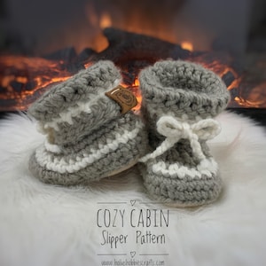 Puede incluir: Patrón de zapatillas Cozy Cabin con un par de zapatillas de crochet grises y blancas con un lazo en una de las zapatillas. Las zapatillas están sobre una superficie blanca y esponjosa frente a una chimenea.