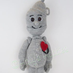 Puede incluir: Un muñeco de hojalata de crochet gris con un corazón rojo en el pecho. El muñeco tiene una cara sonriente con ojos marrones y una boca negra.