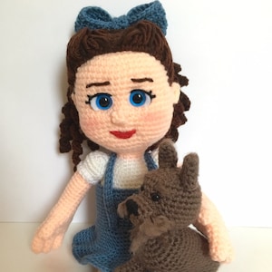 May include: A crocheted doll with brown hair and a blue bow, wearing a blue dress and holding a brown crocheted dog.