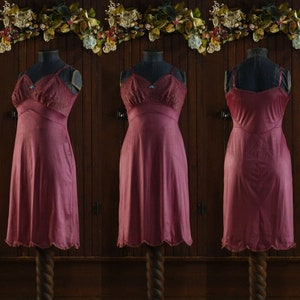 Könnte beinhalten: Ein burgunderfarbenes Schlupfkleid mit Spitzenbesatz und einem gewellten Saum. Das Kleid wird auf einer Schaufensterpuppe vor einem hölzernen Hintergrund präsentiert.