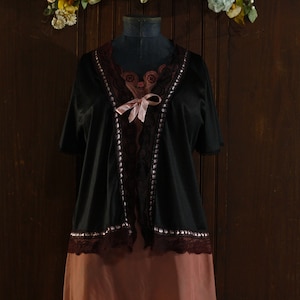 Pode incluir: Uma bata de cetim preto com guarnição de renda e um laço de cetim rosa. A bata é usada sobre um camisão de cetim rosa.