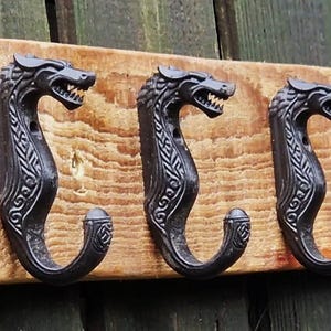 May include: A wooden wall-mounted coat rack with five dark metal dragon-head hooks. The hooks are mounted on a light brown, rectangular wooden board with visible wood grain and a knot. The dragon heads face left.