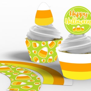 Puede incluir: Dos envoltorios de cupcake de Halloween con un patrón de maíz dulce. Un cupcake está cubierto con un adorno de cupcake de maíz dulce y el otro está cubierto con un círculo verde con el texto "Happy Halloween" y tres adornos de cupcake de maíz dulce. También hay un envoltorio de cupcake de maíz dulce verde y naranja.