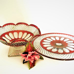 May include: A set of two vintage glass serving dishes with a clear base and a red rim. The dishes have a scalloped edge and a pattern of small diamonds. The larger dish is a bowl with a pedestal base, and the smaller dish is a plate.