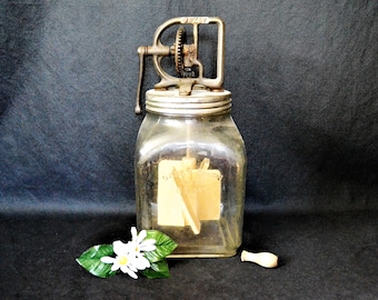 May include: An antique glass butter churn with a metal crank mechanism on top. The jar has a metal lid and a wooden paddle inside. A small wooden stopper and a sprig of white flowers with green leaves are in the foreground.