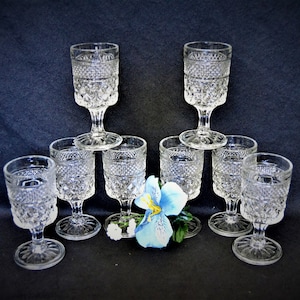 May include: Set of eight clear glass stemmed drinking glasses with a diamond pattern. The glasses are arranged in a pyramid shape with a blue flower in front.