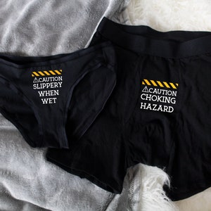 Women's black bikini underwear with phrase "Caution slippery when wet" in white on the front center, men's black boxer brief with "Caution choking hazard" in white on the fly