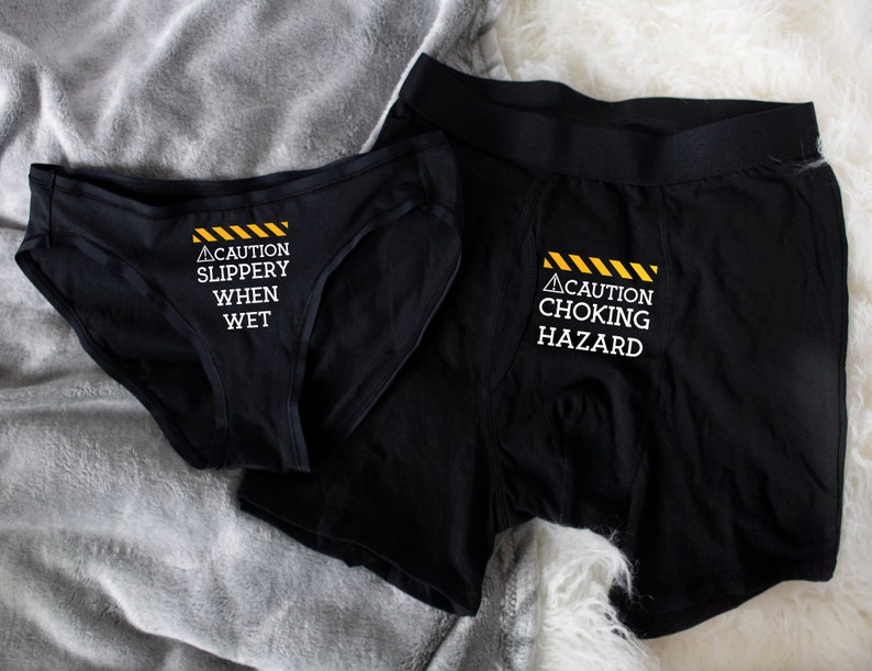 Women's black bikini underwear with phrase "Caution slippery when wet" in white on the front center, men's black boxer brief with "Caution choking hazard" in white on the fly