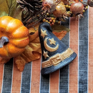 May include: A black and gold witch hat with star and moon details, a small orange pumpkin, and fall foliage. The hat has a gold buckle and belt. The background has orange and white stripes.