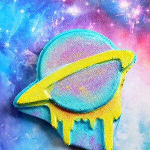 May include: A colorful bath bomb shaped like a planet with a yellow ring and dripping accents. The planet is a mix of blue, purple, and pink with a yellow ring. The background is a vibrant galaxy of blues, purples, and pinks.
